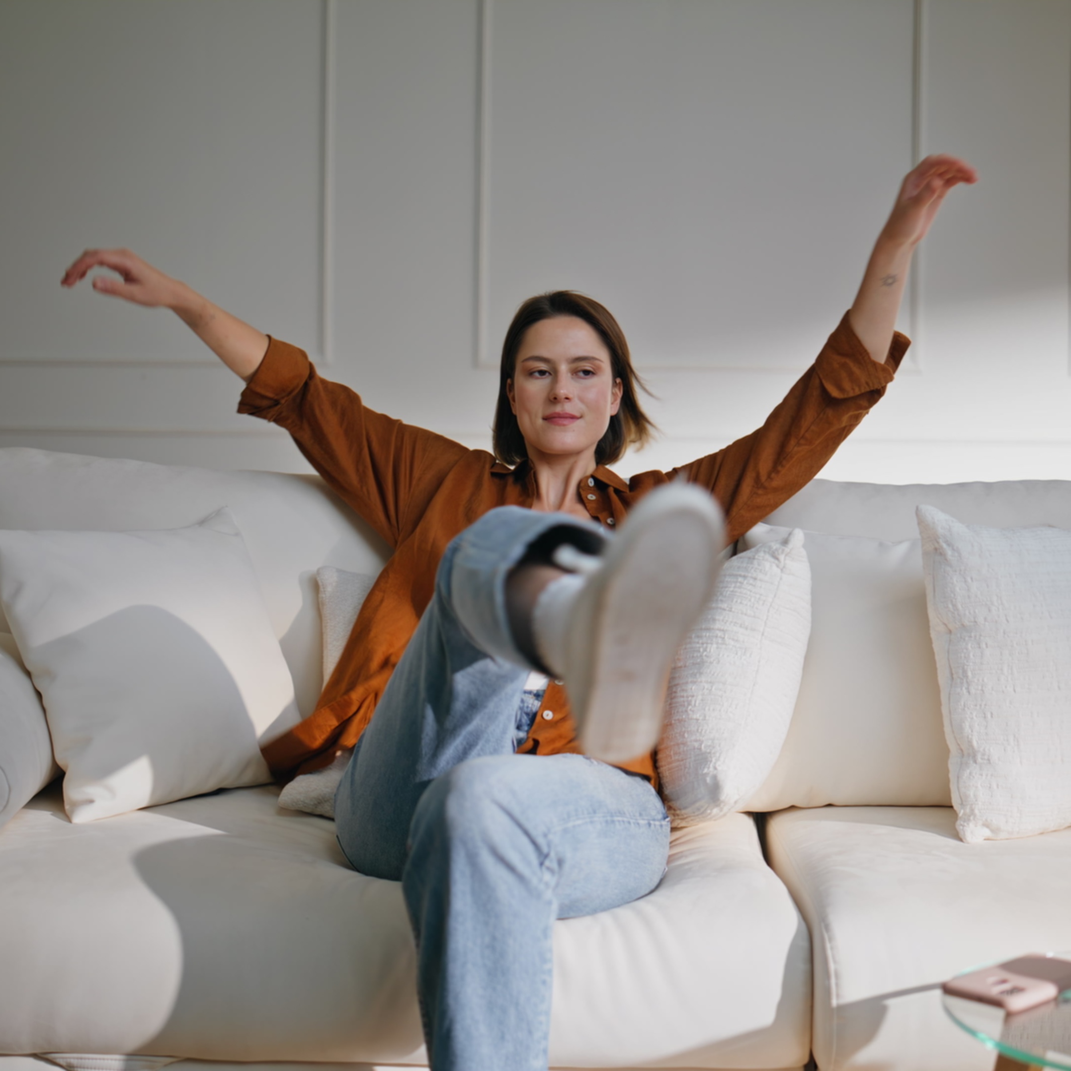 Happy woman lounging on sofa modern living room. Relaxed brunette sit down on cozy couch surrounded by white pillows feeling carefree. Cheerful girl relaxing comfortably in bright home apartment.